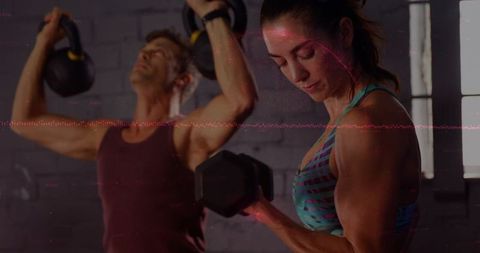Woman Curling Dumbbell While Partner Lifting Kettlebells in Dim Industrial Gym Training