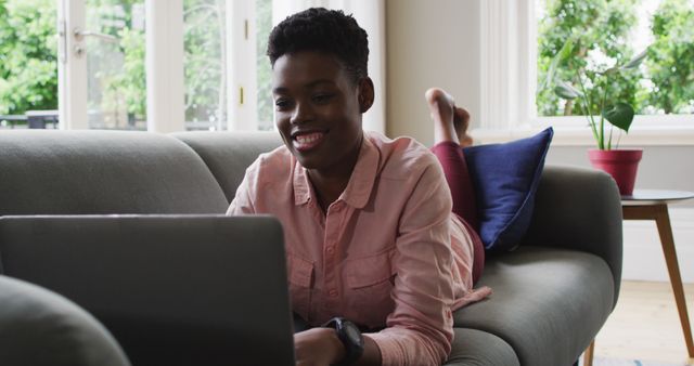 Young African American woman working from home while lying on a couch. She is using her laptop in a bright and comfortable living room with modern decor. This image is perfect for illustrating themes of remote work, freelance lifestyle, productivity, and modern technology usage. Ideal for websites, blogs, social media, and advertisements that focus on working from home, work-life balance, and technology.