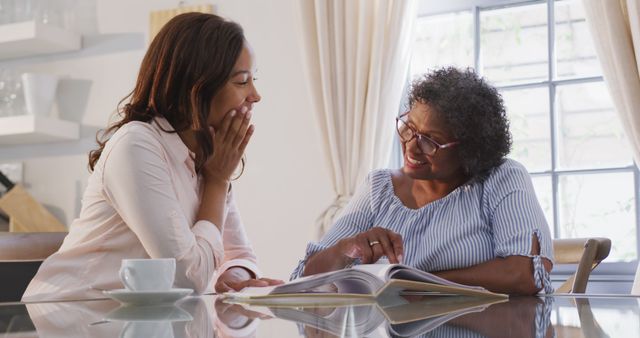 Happy senior african american woman and adult daughter sitting at table and looking at photo album. Lifestyle, free time, family and domestic life.