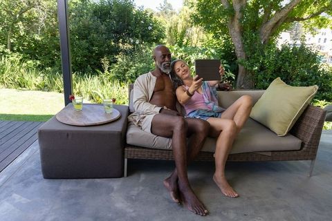 Senior interracial couple relaxing on patio daybed sharing tablet while sipping cocktails