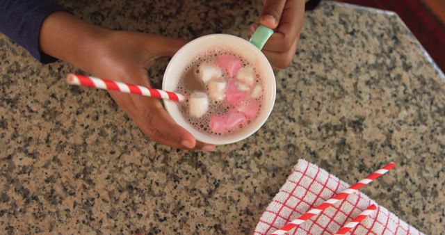 A close-up of a child's hands holding a mug of hot chocolate topped with marshmallows and a red-and-white striped straw. The granite countertop and checkered towel enhance the cozy and inviting atmosphere. Ideal for use in lifestyle, food and drink, or family-oriented materials, particularly in content related to winter, comfort foods, and family moments.