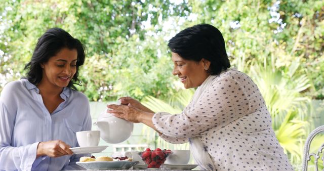 Elderly woman pouring tea for smiling daughter in garden. Ideal for use in family, bonding, or hospitality themes. Can be used for advertisement promoting afternoon tea experiences, familial love and connections, and outdoor activities.