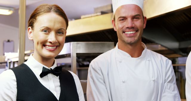 Smiling waitress in black uniform with bow tie standing beside smiling chef in white kitchen attire in restaurant kitchen. Can be used for hospitality industry promotions, teamwork importance, professional culinary environments, restaurant staff training materials.