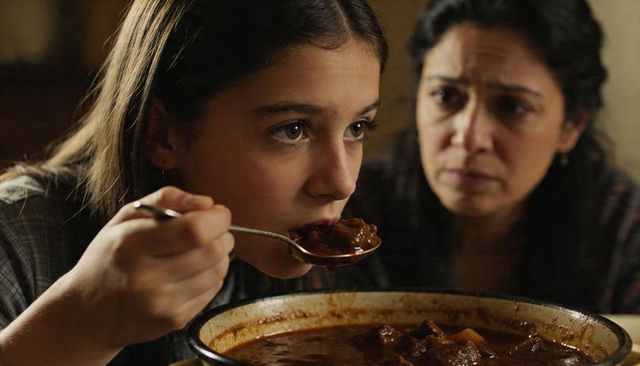 Teen girl tasting hearty stew at kitchen table, watchful mother in background