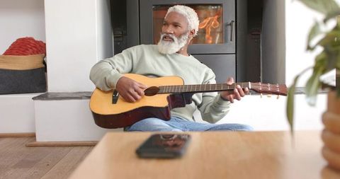 Senior Man Playing Guitar Near Fireplace Relaxing with Smartphone