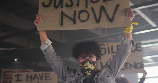 Passionate young adult holding 'Justice Now' sign during a protest, conveying message of advocacy and determination. Wearing a bandana and surrounded by similar signs addressing social issues. Ideal for use in articles, campaigns, or content related to social justice, civil rights movements, activism, and community engagement.