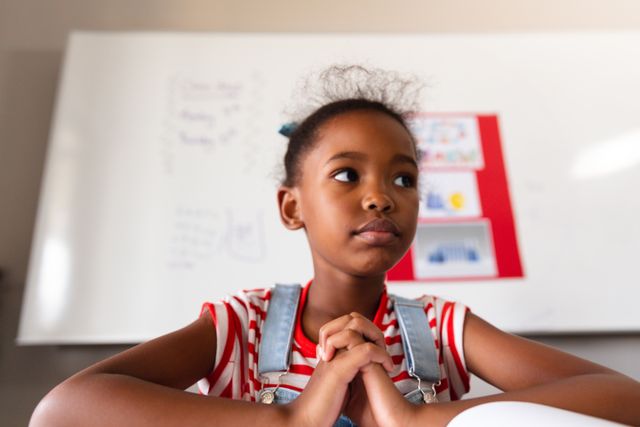 Young African American schoolgirl sitting at desk in classroom, looking thoughtful with hands clasped. Ideal for educational materials, school brochures, learning resources, and articles on childhood education and student life.