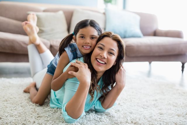 Smiling mother and daughter lying on soft carpet in living room, creating a warm and affectionate atmosphere. Perfect for use in family-oriented advertisements, parenting blog posts, and lifestyle magazines focusing on family relationships and home comfort.