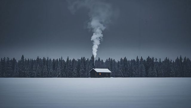 Solitary cabin emitting smoke over snowfield with warm glowing window and pine forest