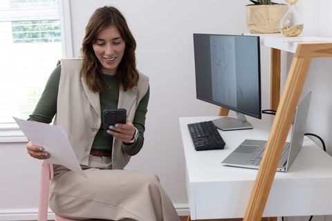 Woman working from home checking smartphone while holding document in minimalist office
