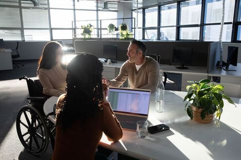 Diverse team collaborating with wheelchair user reviewing spreadsheet at laptop in sunlit office