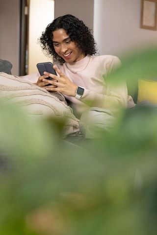 Non-binary person smiling while browsing smartphone on cozy sofa with plants and cushions