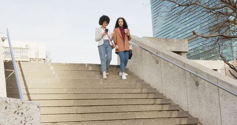 Two women descending urban stairs holding coffee and checking smartphone, city lifestyle