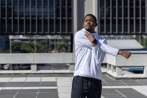 African american man stretching shoulders on urban rooftop wearing white hoodie, smartwatch