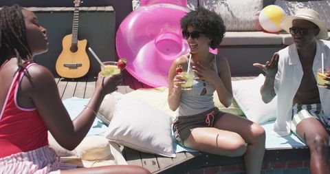 African american friends laughing and sipping cocktails on colorful poolside deck