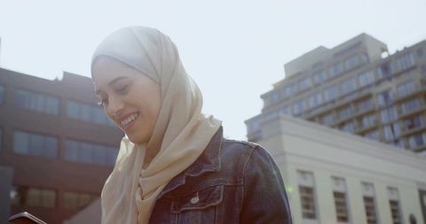 Smiling woman wearing beige hijab and denim jacket checking phone on sunlit city street