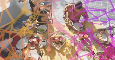 Friends sharing sunlit outdoor feast on yellow tablecloth with roasted bird and passing plates