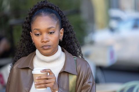 African woman walking on urban sidewalk holding coffee wearing brown leather jacket