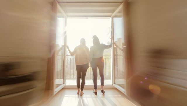 Two women standing at open balcony doors backlit by warm morning sunlight