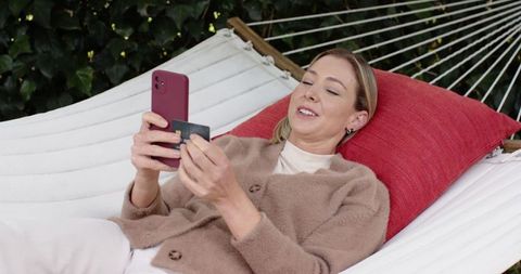 Relaxing woman reclining in hammock using smartphone for online shopping with credit card