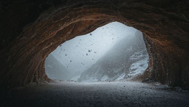Framing snowfall through cave arch revealing icy ridge, wind-blown snow and rust rock