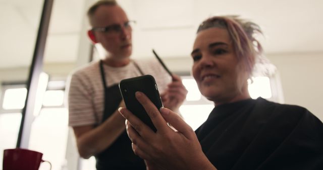 Woman sits in salon chair while hair stylist works on her hair. She is smiling and using a smartphone. Perfect for concepts related to beauty services, customer satisfaction, modern communication, and relaxation. Use this photo on websites and ads for salons, beauty services, mobile apps, and tech companies focusing on connectivity and customer engagement.