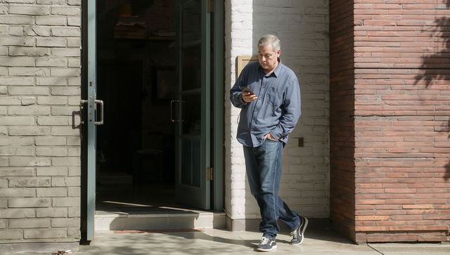 Middle-aged man leaning at open doorway checking smartphone on sunlit urban sidewalk