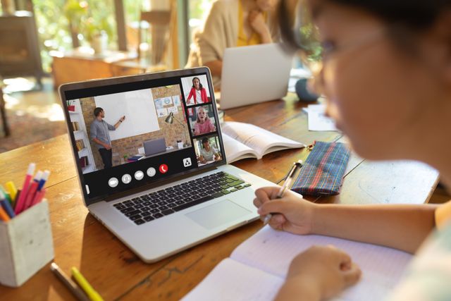 Asian teenage girl writing notes while male teacher teaching over video call in online class at home. Laptop, screen, unaltered, childhood, wireless technology, education, student and e-learning.