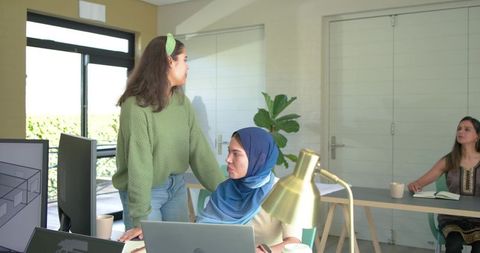 Collaborating women in bright open-plan office, diverse team using laptops and monitors