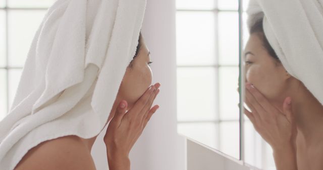 Young woman engaging in her skincare routine with a towel wrapped on her head. Suitable for promoting beauty and skincare products, self-care blogs, and morning routine tips.