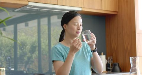 Asian woman taking daily medication with glass of water in bright modern kitchen