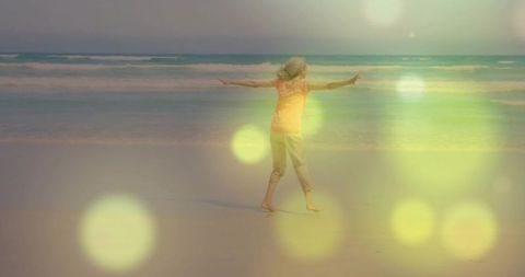 Senior woman walking barefoot along sunlit beach with bokeh lens flare