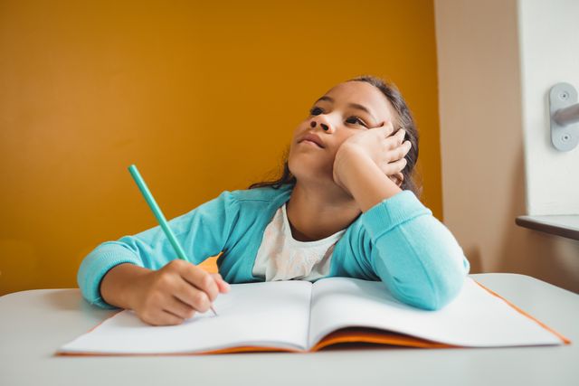Young girl sitting at desk in classroom, holding pencil and writing in notebook while daydreaming. Ideal for educational content, school-related articles, and materials focusing on childhood learning and imagination.