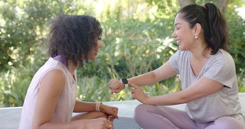 Multiracial women comparing smartwatch after outdoor yoga, smiling and hydrating