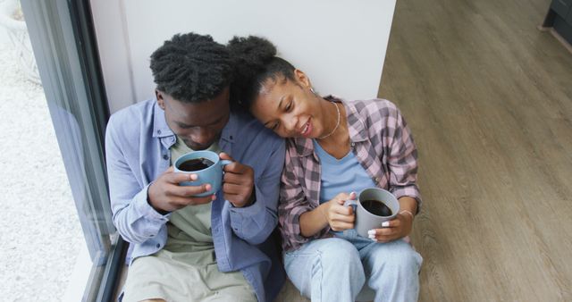 Couple sitting on floor against a wall, drinking coffee, and smiling. Suggests intimacy, connection, and comfort. Ideal for use in lifestyle blogs, relationship advice articles, coffee product advertisements, and indoor lifestyle presentations.