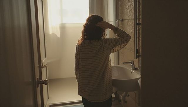 Backlit woman touching hair in home bathroom by frosted window at sunrise