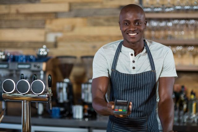 Smiling barista holding a credit card reader in a modern cafe. Ideal for illustrating themes related to customer service, small business, technology in hospitality, and modern payment methods. Suitable for use in articles, advertisements, and websites focused on cafes, restaurants, and customer interactions.