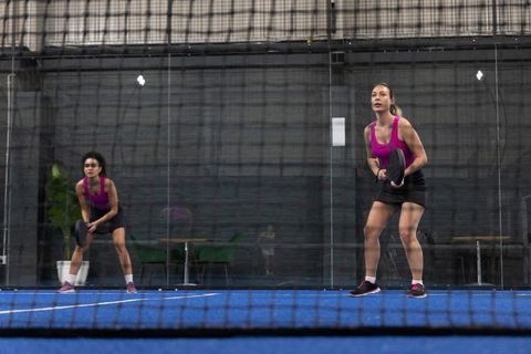 Women playing padel doubles in magenta tanks on indoor blue court with glass walls and net