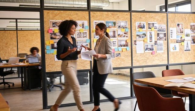 Walking colleagues discussing project in modern glass office with sticky notes, teamwork