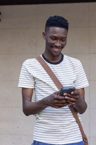 Young african american man smiling while texting on smartphone with crossbody bag