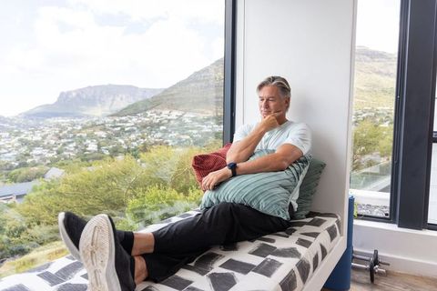 Mature man relaxing on window bench, contemplating mountain view with panoramic landscape
