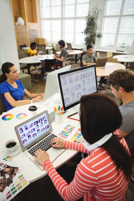 Image shows a group of graphic designers working together in a contemporary office environment. They are using computers and laptops, discussing color palettes, and brainstorming ideas. Ideal for illustrating concepts of teamwork, creativity, and modern workspaces in marketing materials, business presentations, or website content.