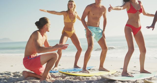 Group of young friends enjoying learning to surf on a sunny beach. They are standing and crouching on the sand practicing with surfboards, wearing colorful swimwear, and sharing moments of fun and excitement. Perfect for use in advertisements, articles about summer activities, beach tourism, lifestyle blogs, and wellness content.
