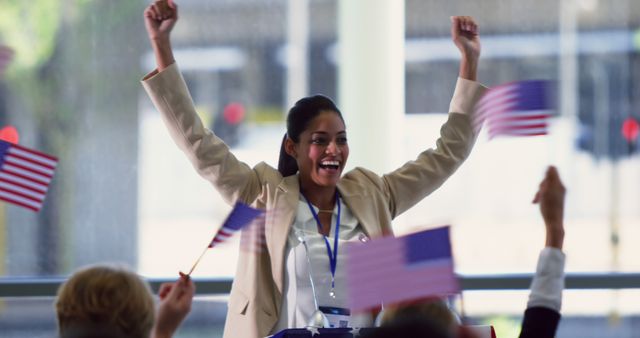 Woman is expressing joy and enthusiasm in a political event setting, raising her arms while holding American flags. This image can be used for political campaigns, motivational speaking events, female leadership, patriotism-themed projects, and celebratory occasions.