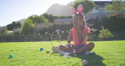 Young girl wearing bunny ears sitting on lawn enjoying easter egg hunt and candy in sunny backyard