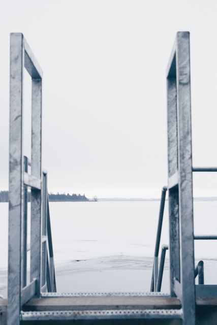 Metal dock leading to a frozen lake with a snow-covered landscape. Perfect for themes of winter, solitude, cold environments, and nature. Useful for winter travel brochures, nature magazine covers, and outdoor adventure promotions.
