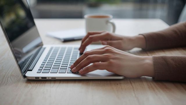 Hands typing on laptop with coffee and notebook in sunlit minimal workspace