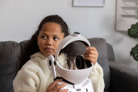 Curious child holding futuristic white headset and home robot on cozy gray couch
