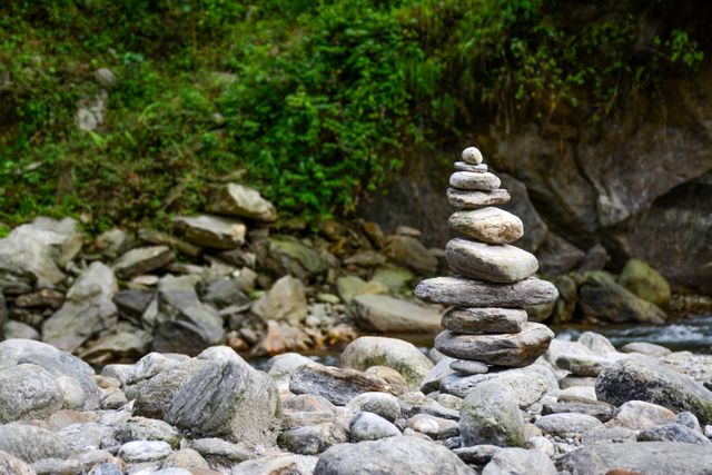 Balanced stone cairn in a serene mountain riverbed with lush green vegetation in the background. Ideal for themes related to balance, mindfulness, meditation, and nature. Perfect for use in wellness and relaxation content, environmental campaigns, outdoor activities promotions, and travel blogs.