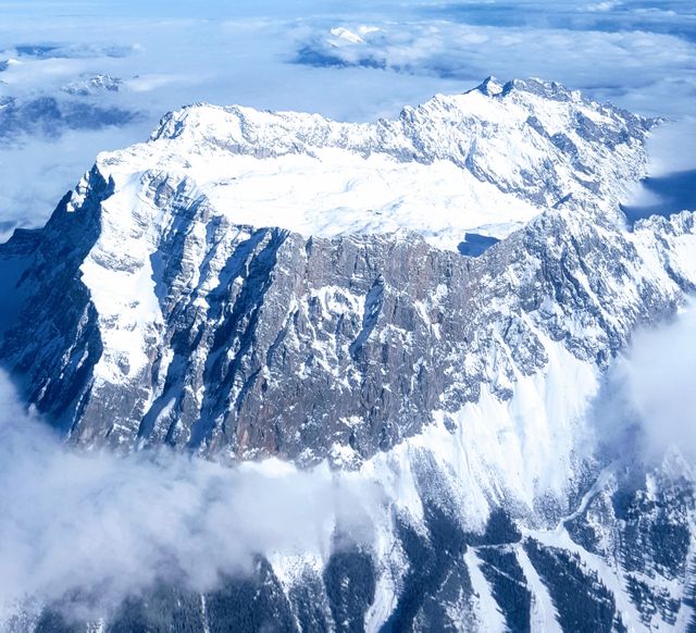 This image shows an aerial view of snow-capped mountain peaks towering above the clouds in a winter landscape. The rugged and majestic terrain makes it ideal for depicting adventure, travel, nature, and outdoor activities. It can be used in travel brochures, environmental conservation campaigns, wallpaper backgrounds, and winter vacation promotions.